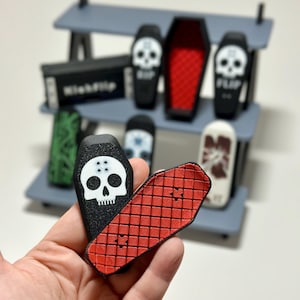 May include: A hand holding a black coffin-shaped fingerboard with a white skull graphic and a red fingerboard with a grid pattern. Other fingerboards and a shelf are in the background.