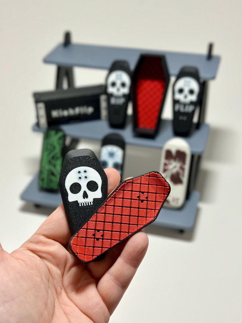 May include: A hand holding a black coffin-shaped fingerboard with a white skull graphic and a red fingerboard with a grid pattern. Other fingerboards and a shelf are in the background.