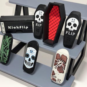 May include: Miniature black skateboards with various designs, including a circuit board, sugar skull, and floral pattern, displayed on a gray tiered shelf. A black coffin and a box with "Kickflip" are also present.
