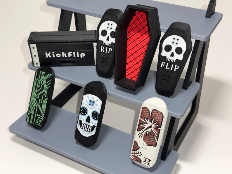 May include: Miniature black skateboards with various designs, including a circuit board, sugar skull, and floral pattern, displayed on a gray tiered shelf. A black coffin and a box with "Kickflip" are also present.