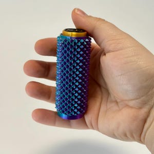 Puede incluir: Un objeto cil&iacute;ndrico con una superficie texturizada de p&uacute;as iridiscentes azules, moradas y turquesas. La parte superior tiene una banda dorada y un centro negro. El objeto se sostiene en una mano sobre un fondo blanco.