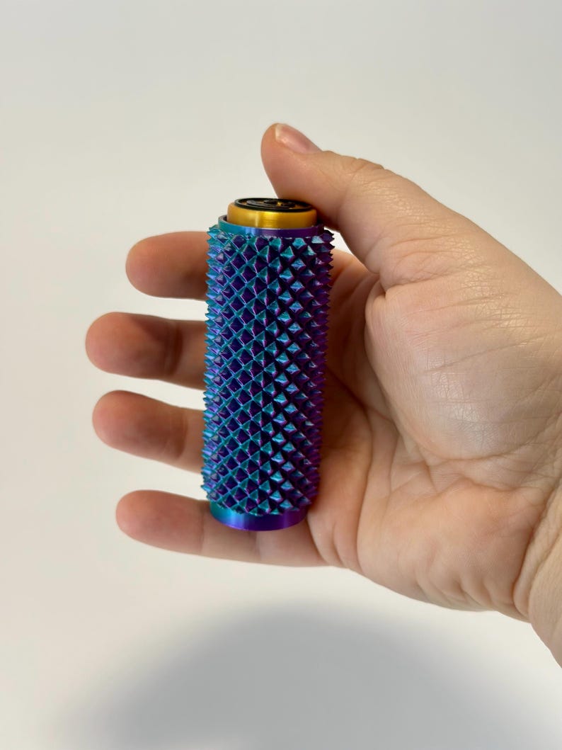 Puede incluir: Un objeto cil&iacute;ndrico con una superficie texturizada de p&uacute;as iridiscentes azules, moradas y turquesas. La parte superior tiene una banda dorada y un centro negro. El objeto se sostiene en una mano sobre un fondo blanco.