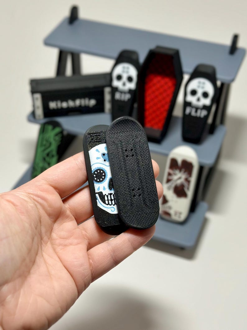 May include: Miniature finger skateboards with skull and coffin designs. One board features a sugar skull design with blue and white accents. Other boards have RIP and FLIP text. The display shelf is gray.