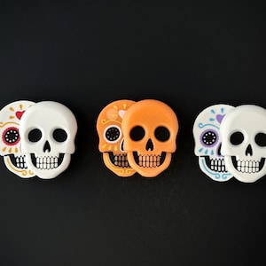 May include: Three pairs of decorative skull figurines in white, orange, and decorated with colourful patterns. The skulls are arranged on a black surface, ideal for Halloween or Day of the Dead decorations.