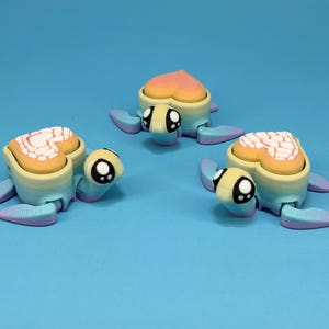 May include: Three whimsical turtle figurines with heart-shaped shells in shades of yellow, orange, and white. The turtles have blue, purple, and yellow bodies and flippers, and large, expressive eyes. The background is a solid light blue.