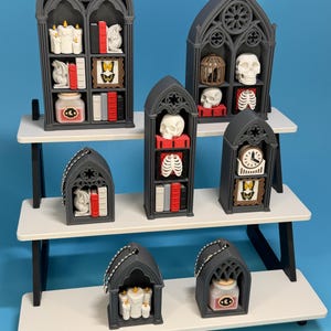 May include: A display of miniature gothic-style shelves, each filled with themed decor. The shelves are dark gray and arranged on a white tiered stand. Items include candles, books, skulls, and framed pictures, creating a spooky, decorative theme.