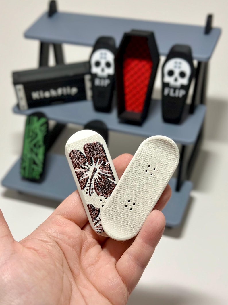 May include: Miniature finger skateboards, one with a brown floral design and the other plain. The set includes a gray shelf with miniature coffins, a box labeled "Kickflip", and a green skateboard.