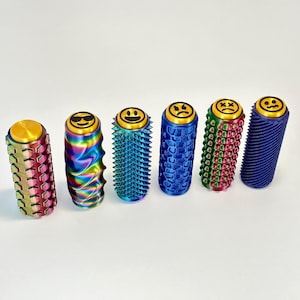 Puede incluir: Seis coloridos juguetes fidget cil&iacute;ndricos con dise&ntilde;os de caras sonrientes en la parte superior. Los juguetes presentan varias texturas y colores, incluyendo rosa, arco&iacute;ris, azul, verde y morado. Cada juguete mide unos 7,6 cm de alto.