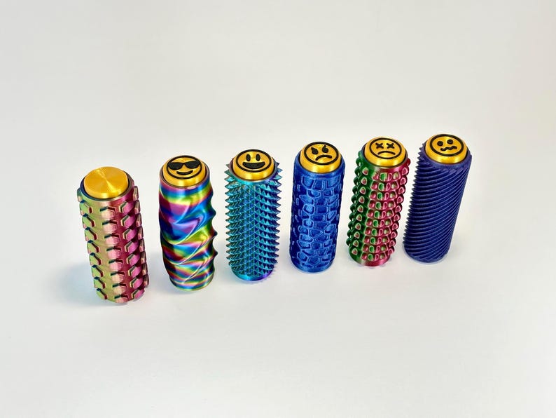 Puede incluir: Seis coloridos juguetes fidget cil&iacute;ndricos con dise&ntilde;os de caras sonrientes en la parte superior. Los juguetes presentan varias texturas y colores, incluyendo rosa, arco&iacute;ris, azul, verde y morado. Cada juguete mide unos 7,6 cm de alto.