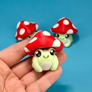 May include: Three whimsical figurines of a frog wearing a red mushroom cap with white spots. The frog is light green with a white belly and large black eyes. The background is a solid blue.