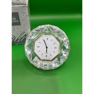 Puede incluir: Un reloj de escritorio de cuarzo transparente y facetado con esfera blanca y manecillas negras. El reloj tiene un bisel dorado y la palabra "QUARTZ" está impresa en la esfera. El reloj está junto a una caja con las palabras "TIME CLOCK" impresas.
