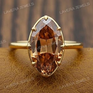 Op de afbeelding: Een gouden ring met een grote, marquise-geslepen, cognac-kleurige edelsteen. De ring heeft een dunne, gepolijste gouden band en de edelsteen is gezet in een delicate gouden bezel. De ring wordt gepresenteerd op een bruin leren oppervlak. De tekst "LAVERA JEWELS" is zichtbaar.
