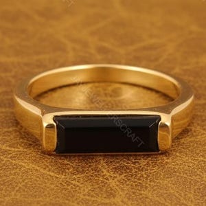 Könnte beinhalten: Ein goldfarbener Ring mit einem rechteckigen schwarzen Stein in der Mitte. Der Ring hat ein schlichtes, modernes Design und wird auf einer braunen Lederoberfläche präsentiert. Der Ring ist ein Schmuckstück.