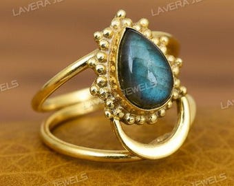 Può includere: Un anello color oro con una pietra preziosa blu-verde a forma di goccia. L'anello ha un design a doppia fascia con una forma a mezzaluna. La pietra preziosa è circondata da un bordo di perline.