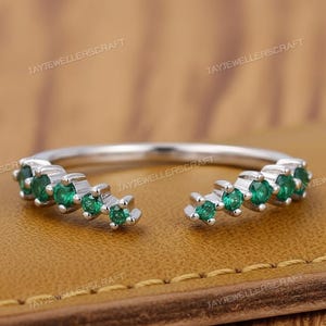 Peut inclure: Une bague en argent avec un design à tige fendue, ornée d'une série de petites pierres précieuses vertes vives. La bague est posée sur une surface en cuir marron, mettant en valeur les détails du bijou.