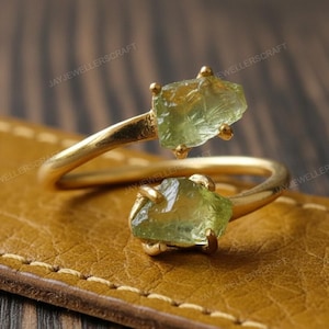 Puede incluir: Un anillo dorado con dos piedras preciosas en bruto de color verde claro. La banda del anillo se curva alrededor del dedo, con una piedra preciosa en cada extremo. El anillo está sobre una superficie de cuero marrón.