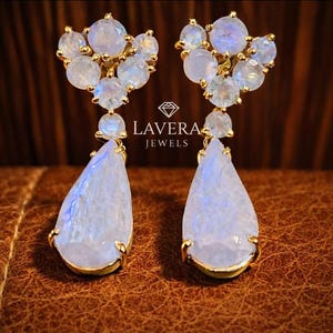 May include: A pair of gold earrings with light blue gemstones. The earrings have a cluster of small, round stones at the top and a larger, teardrop-shaped stone below. The brand name, Lavera Jewels, is visible.