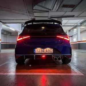 May include: A dark blue hatchback car viewed from the rear, featuring sharp red LED taillights, a black roof spoiler, and a dual-tip exhaust system. The number plate reads 1234 ABC, and a small red N emblem is visible above the rear diffuser.