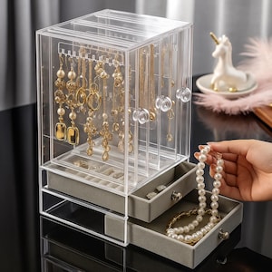 May include: Clear acrylic jewellery organiser with multiple compartments and drawers. The organiser displays various gold necklaces, earrings, and bracelets. The drawers are lined with grey fabric. A hand holds a pearl necklace.