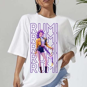 Puede incluir: Camiseta blanca con un gráfico de una mujer con chaqueta amarilla y shorts de mezclilla, con el nombre "RUMI" en texto vertical morado. La mujer tiene el pelo morado y sostiene un arma. La camiseta tiene mangas cortas.