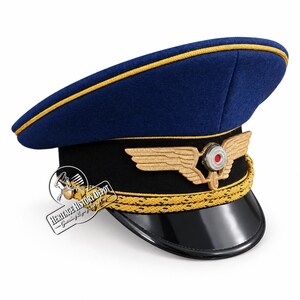 May include: A blue peaked cap with a black band and a shiny black visor. The cap features gold piping along the crown, a gold braided chin strap, and a gold-toned winged emblem with a central red and white circular detail on the front band.