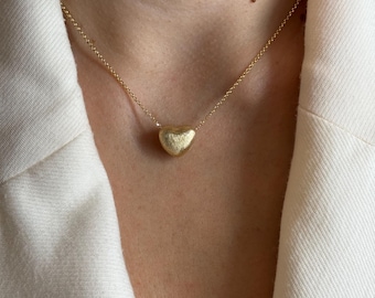 Può includere: Una collana color oro con un ciondolo a forma di cuore. Il ciondolo ha una superficie strutturata ed è appeso a una catena delicata. La collana è mostrata su uno sfondo bianco.