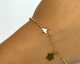 May include: A delicate gold-colored bracelet with a small cross pendant and a clover charm. The bracelet is made of a fine chain and is worn on a wrist. The cross pendant has a white enamel filling. The clover charm hangs freely from the chain.