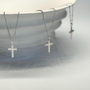 May include: A collection of silver cross necklaces, each with a small, polished cross pendant suspended from a fine chain. The necklaces are arranged against a white backdrop, creating a minimalist display. The jewellery is likely made of stainless steel, and suitable for everyday wear.