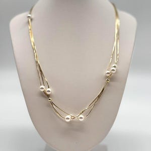 Könnte beinhalten: Eine goldfarbene Halskette mit mehreren feinen Strängen und kleinen, runden, weißen Perlen. Die Halskette wird auf einer weißen Schaufensterpuppe präsentiert, die ihr mehrschichtiges Design und ihre filigranen Details hervorhebt. Die Perlen sind gleichmäßig über die Stränge verteilt.
