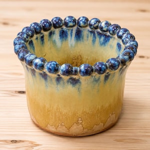 Peut inclure: Un pot en céramique doté d'un bord perlé avec un vernis bleu profond. Le corps du récipient passe d'un motif goutte à goutte bleu vif sur le dessus à une teinte jaune doré chaude à la base, présentant une finition émaillée lisse sur l'ensemble.