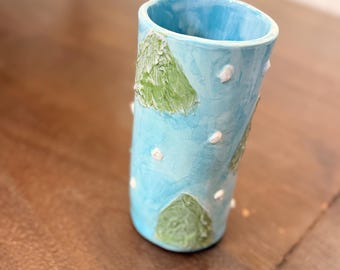 Può includere: Un bicchiere in ceramica fatto a mano con esterno azzurro chiaro, con forme verdi strutturate a forma di montagna e piccoli puntini bianchi. L'interno è di una tonalità di blu più scura. Il bicchiere è cilindrico.