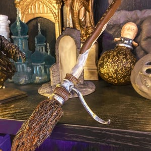 Op de afbeelding: Een gedetailleerde miniatuur bezemsteel met een bruine steel en een geweven, takachtig hoofd. De steel heeft een zilverkleurige metalen accent. Het wordt tentoongesteld op een plank met andere fantasie-thema figuren.