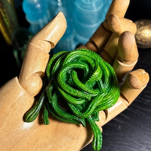 Puede incluir: Una escultura verde vibrante y de diseño intrincado que se asemeja a una planta enrollada o un tentáculo, sostenida en una mano de madera. La escultura tiene una superficie texturizada y está colocada sobre un fondo oscuro. Otras estructuras azules translúcidas son visibles en el fondo.