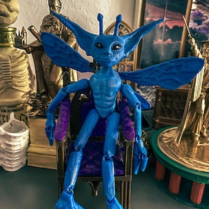 May include: A blue articulated figurine of a creature with large ears, wings, and insect-like features, seated on a miniature chair with purple cushions. The figurine has a detailed, textured surface and is posed with its arms and legs bent.