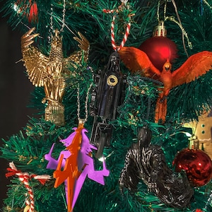 Pode incluir: Uma árvore de Natal decorada com vários enfeites. Enfeites de águia dourada, figura preta, fénix laranja e bola vermelha são visíveis. A árvore é verde com luzes e outros elementos decorativos.