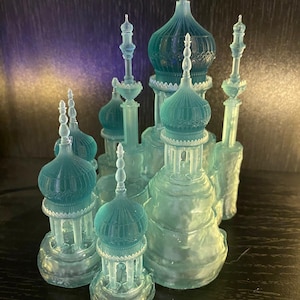 Puede incluir: Un castillo en miniatura translúcido de color verde azulado con múltiples cúpulas y agujas. El modelo arquitectónico tiene un diseño detallado con patrones intrincados y un acabado brillante. El castillo se exhibe sobre una superficie oscura, mostrando su delicada estructura y atractivo artístico.