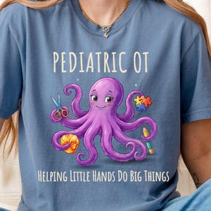 May include: A blue t-shirt featuring a purple octopus illustration holding scissors, a yellow object, a pencil, and a puzzle piece. The text reads PEDIATRIC OT and HELPING LITTLE HANDS DO BIG THINGS. The design is centered on the front of the shirt.