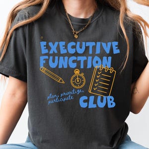 May include: A charcoal gray t-shirt featuring the blue text "EXECUTIVE FUNCTION" above a yellow pencil, stopwatch, and checklist icon, with the blue text "plan, prioritize, participate" and "CLUB" printed below the graphic elements.