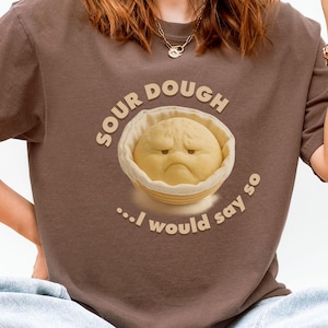 May include: A brown t-shirt featuring a cartoon sourdough bread character with a grumpy expression inside a bowl. The text "SOUR DOUGH ...I would say so" is arched above and below the image. The shirt is worn by a person.