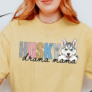 May include: A yellow short-sleeved t-shirt featuring a graphic of a Siberian Husky dog. The text HUSKY is printed in colorful block letters above the script text drama mama. The design includes a detailed illustration of a husky face.
