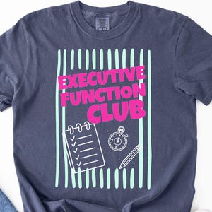 May include: A dark blue short-sleeve t-shirt featuring a graphic print with vertical light green stripes. The text "EXECUTIVE FUNCTION CLUB" is printed in bright pink, accompanied by white line art of a checklist, a stopwatch, and a pencil.