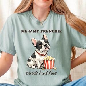 以下が含まれることがあります： セージグリーンの半袖Tシャツ。赤い首輪をした白黒のフレンチブルドッグと、赤と白のストライプのポップコーンバケツのグラフィックが描かれています。ダークブラウンの文字で「ME & MY FRENCHIE」および「snack buddies」とプリントされています。