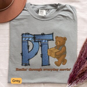 Peut inclure: T-shirt gris avec un motif graphique "PT" en jean et un ours en peluche brun tenant une botte de foin. Le texte "Healin' through everyday movin'" est imprimé en dessous. L'étiquette indique "Grey".