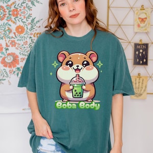 May include: Teal-colored t-shirt featuring a cartoon hamster holding a boba drink. The hamster has a brown body, pink cheeks, and large eyes. The text "Boba Body" is printed in green below the hamster. The shirt is a relaxed fit.