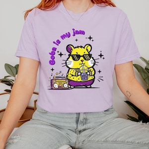 May include: A light purple t-shirt featuring a cartoon hamster wearing sunglasses and holding a boba drink. The text "Boba is my jam" is arched above the hamster. A retro boombox is to the left of the hamster.