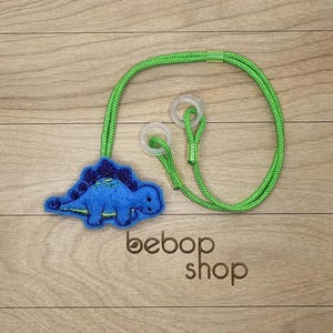May include: A blue dinosaur-shaped felt item with a green cord and clear plastic loops. The dinosaur has a dark blue spine and is attached to a bright green cord. The text "bebop shop" is printed on the wooden surface.