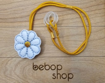 Puede incluir: Una margarita de fieltro blanco con un centro amarillo y costuras azules cuelga de un cordón amarillo con un cierre de plástico transparente. El texto "bebob shop" está impreso en una superficie de madera.