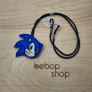 May include: A blue and white embroidered Sonic the Hedgehog character charm attached to a black lanyard with clear plastic adjustable stoppers. The lanyard is on a wooden surface with the text "bebob shop" printed on it.