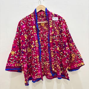 Peut inclure: Veste ouverte magenta vif, ornée de broderies florales complexes jaunes, roses et vertes. La veste présente des bordures bleues et l'étiquette indique "F32".