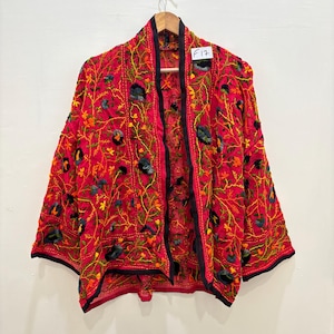 Könnte beinhalten: Eine leuchtend rote Jacke mit aufwendiger Blumenstickerei in Gelb-, Grün- und Schwarztönen. Die Jacke hat einen schwarzen Besatz und hängt an einem Holzbügel. Das Etikett lautet "F17".